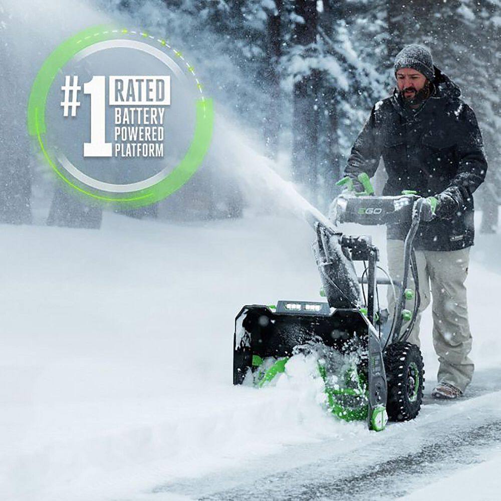 Man using battery powered snow blower on snowy driveway in winter forest setting