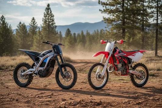 Electric dirt bike and gas dirt bike parked outdoors on a dirt trail with trees and mountains