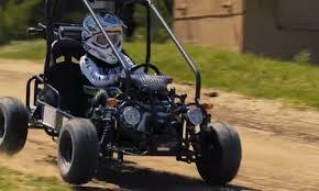 Child riding a gas-powered go-kart on a dirt track outdoors, wearing a helmet and gear.