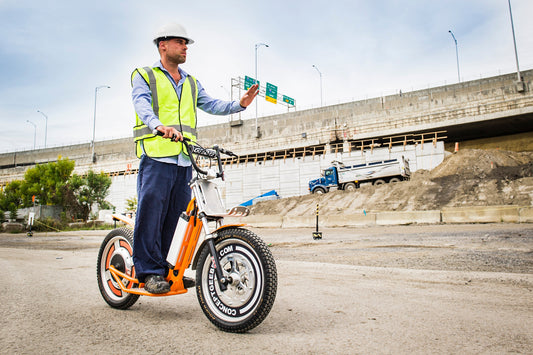 Construction worker on electric scooter near highway, wearing safety vest and helmet
