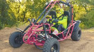 Two people in safety helmets riding a red off-road go-kart on a dirt trail outdoors