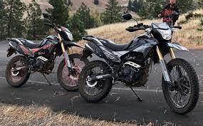 Two dual sport dirt bikes parked on a paved road with hills and trees in the background