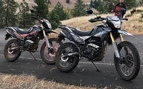 Two off-road dirt bikes with rugged tires parked on a paved road in a scenic outdoor setting