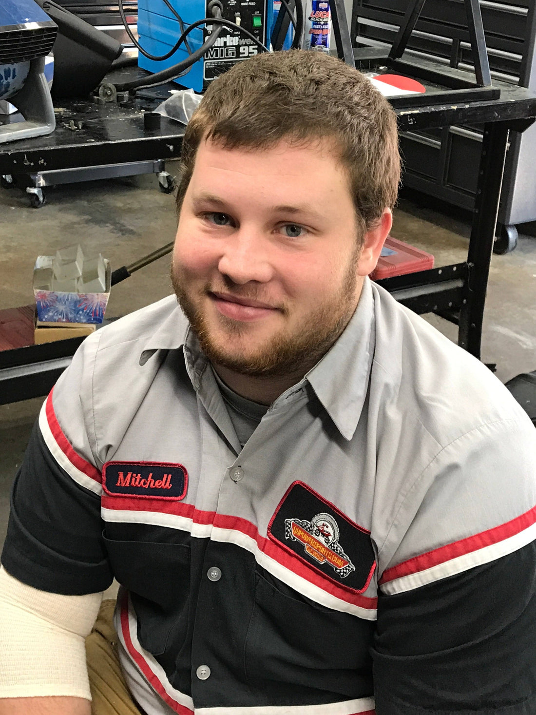 Mechanic in uniform at scooter and moped repair shop with tools and equipment in background.
