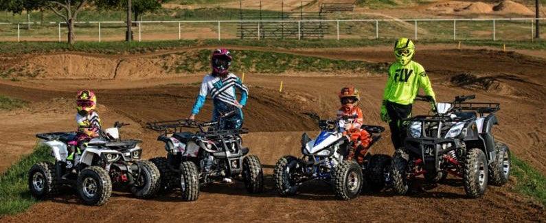 Group of youth and adult riders with ATVs on a dirt track, wearing helmets and motocross gear