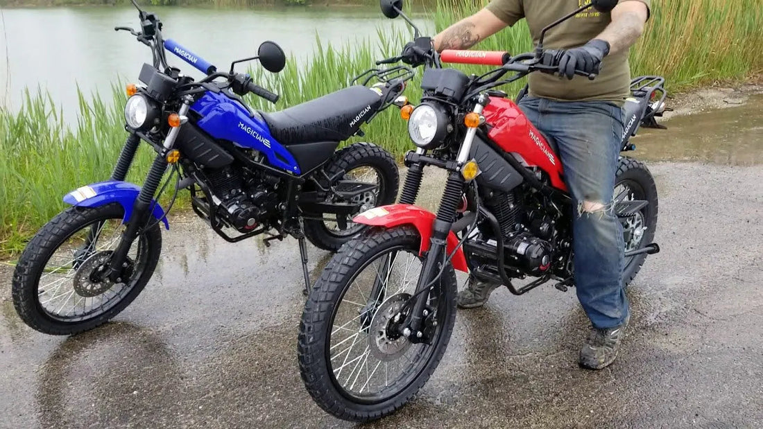Two Magician brand dirt bikes, one blue and one red, parked on wet pavement near a lake with a person sitting on the red bike