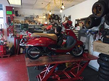 Mechanic servicing red scooter in a workshop with tools and spare tires.