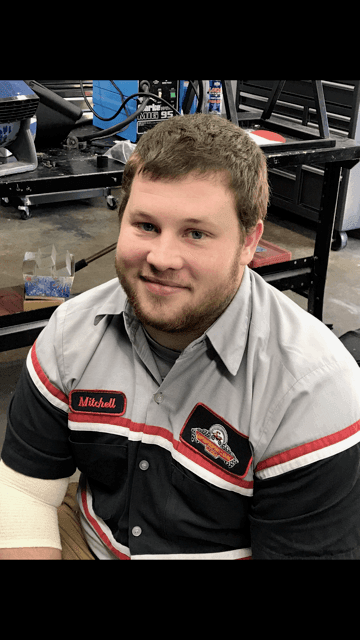 Mechanic in gray uniform with name patch seated in an ATV and dirt bike workshop