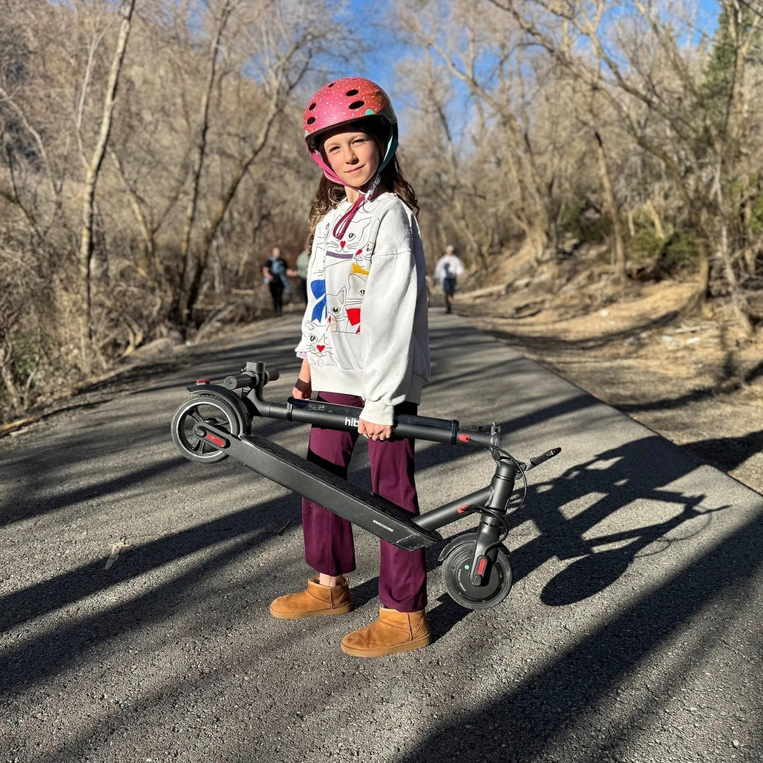 Child with pink helmet holding folded electric scooter on a paved trail outdoors