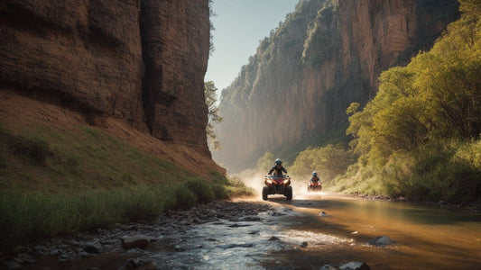 Two riders on ATVs driving along a scenic river trail between tall cliffs and lush trees.