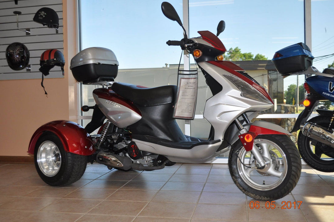 Red and silver 3-wheeled trike motor scooter with trunk, displayed indoors near helmets