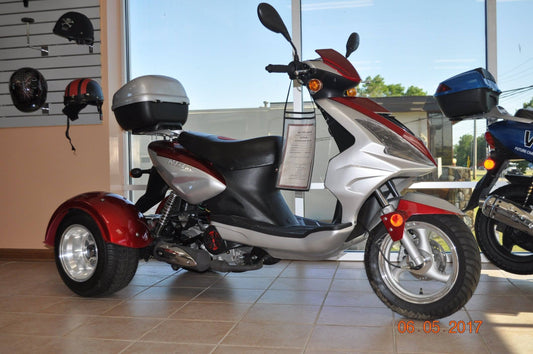 Red and silver 3-wheeled trike motor scooter with trunk, displayed indoors near helmets