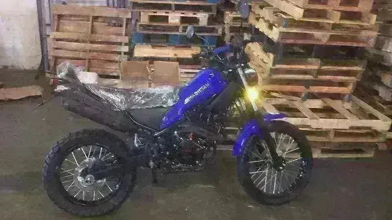 Blue 250cc enduro dirt bike with off-road tires parked indoors by wooden pallets