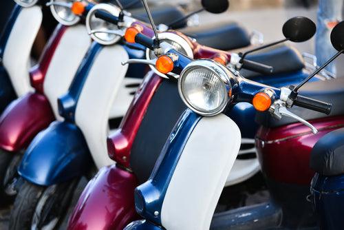 Row of blue and red scooters parked outdoors, close-up of handlebars and headlights.