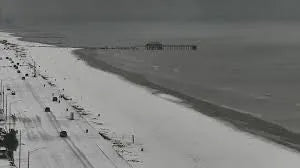 Snow-covered Texas coastline with pier, vehicles on road, and cloudy sky in winter.