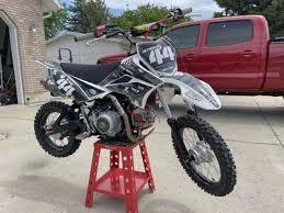 Used dirt bike with black and white graphics on a stand in driveway, red pickup in background
