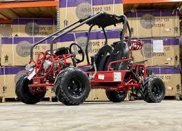Red go-kart with roll cage and off-road tires displayed in warehouse with boxed ATVs